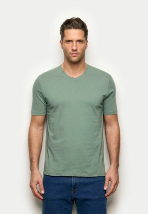 Mann mit kurzen dunklen Haaren, der ein einfaches grünes V-Ausschnitt-T-Shirt und blaue Jeans trägt, steht vor einem weißen Hintergrund.