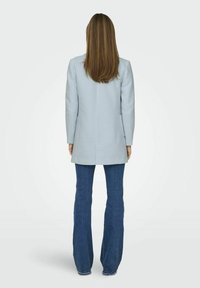 Palton personalizat de culoare albastru deschis, cu o siluetă dreaptă, mâneci lungi și buzunare laterale, asortat cu blugi din denim evazați.