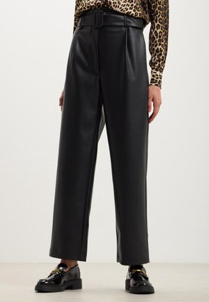 Frau trägt schwarze, hoch taillierte, weite Kunstlederhose mit Gürtel und langärmligem Oberteil mit Leopardenmuster, kombiniert mit schwarzen Loafers.