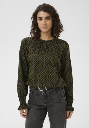 Femme aux cheveux bouclés portant un chemisier vert olive à motifs de feuilles noires et un jean noir taille haute.