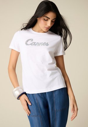 Młoda kobieta o długich ciemnych włosach, ubrana w białą koszulkę z błyszczącym napisem „Cannes” oraz niebieskie dżinsy, pozująca na neutralnym tle.