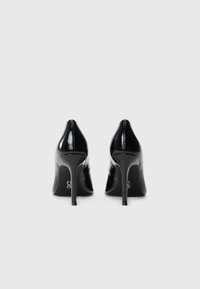 Pair of black patent leather high-heeled shoes viewed from the back on a plain light-gray background.