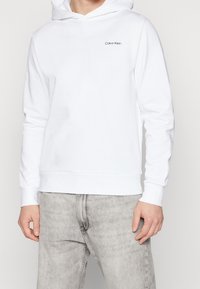 Hoodie bianco realizzato in morbido tessuto, con tasca a marsupio, polsini a costine e un piccolo logo nero di Calvin Klein sul petto.