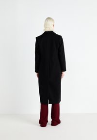 Zwarte lange jas met een rechte snit, notch-kraag en een achterkantventilatie. Gemaakt over rode wijde broek. Minimalistisch ontwerp, geen zichtbare hardware.