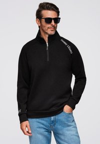 Sudadera de forro polar negra con media cremallera y textura suave. Presenta el texto blanco "OMBRE EXTIMO" en el hombro y puños acanalados. Combinada con jeans azules.