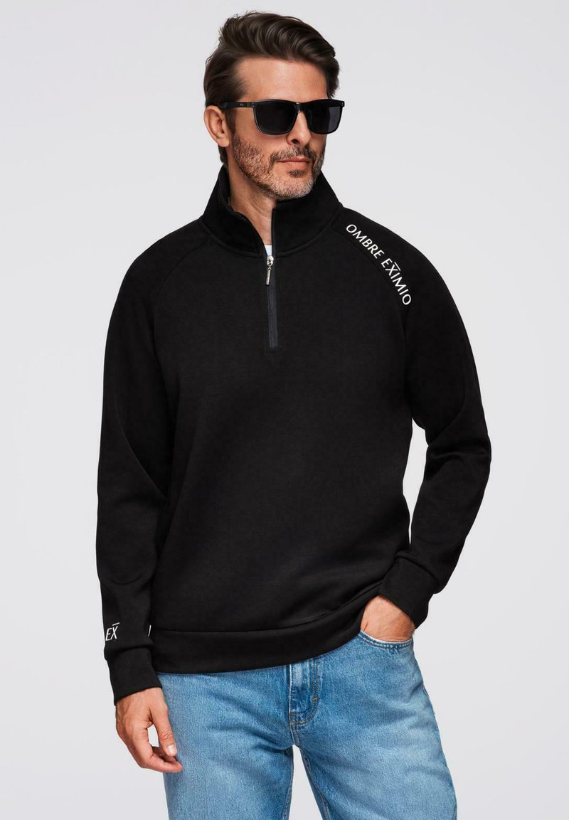 Sudadera de forro polar negra con media cremallera y textura suave. Presenta el texto blanco "OMBRE EXTIMO" en el hombro y puños acanalados. Combinada con jeans azules.