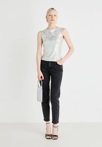 Sølvpaljett top uten ermer, høy hals, kombinert med svarte Skinny-jeans. Modellen holder en liten sølvveske med metallhåndtak. Svarte hælsandaler.