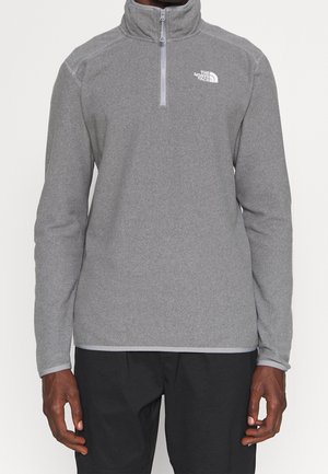 Mann trägt einen grauen Fleecepullover mit Viertel-Reißverschluss und langen Ärmeln sowie schwarze Hose, steht vor schlichtem Hintergrund.