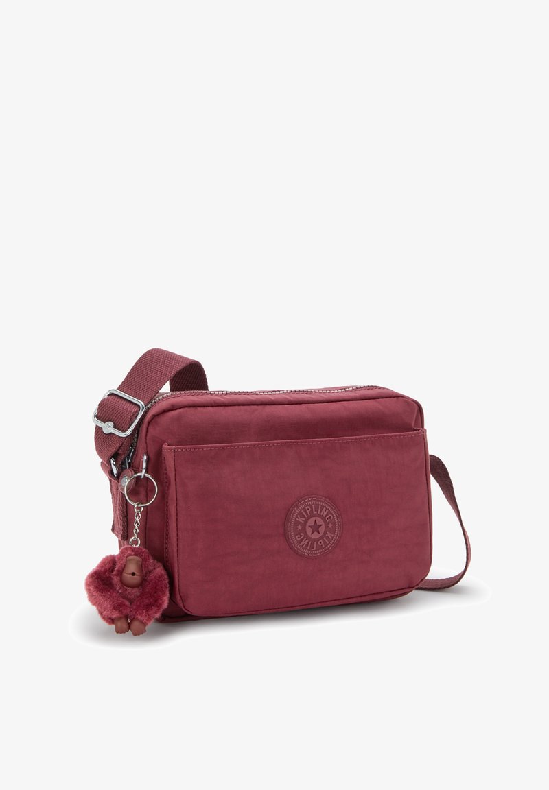 Borsa a spalla in tessuto rosso con chiusura a zip, tasca anteriore e patch logo circolare; presenta un portachiavi a forma di scimmia realizzato in materiale simile alla pelliccia.