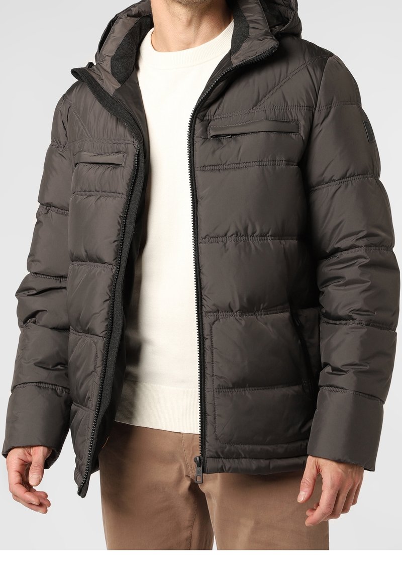 Mies, który ma na sobie ciemno-brązową pikowaną kurtkę z zamkiem błyskawicznym z przodu, kieszeniami na piersi i beżowym swetrem, stoi z luźno splecionymi rękami.