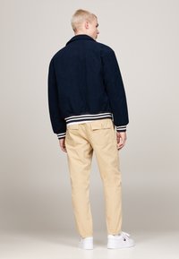 Giacca bomber in suede blu navy con polsini a righe bianche e blu navy, abbinata a chinos beige e sneakers bianche. Sfondo chiaro.