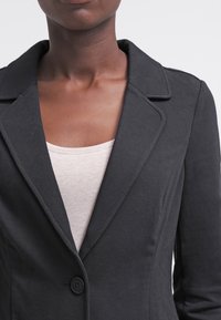 Černý saka na míru s klopovým límcem, jednoduchým zapínáním na knoflík a hladkým materiálem, spojené se světlým, těsně střiženým topem pod ním.