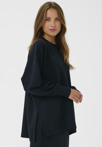 Vrouw met lang lichtbruin haar die een loszittend zwart T-shirt met lange mouwen draagt, staande in profiel tegen een effen witte achtergrond.