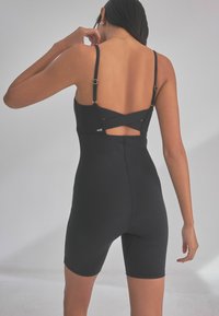 Body nero con spalline regolabili, design incrociato sul retro e vestibilità aderente. Texture liscia con un logo minimalista vicino alla vita.