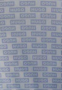 Blå mönstrad tyg med upprepade "Hugo"- och "O7N"-logotyper, arrangerade i ett rutnät på en texturerad vit bakgrund.