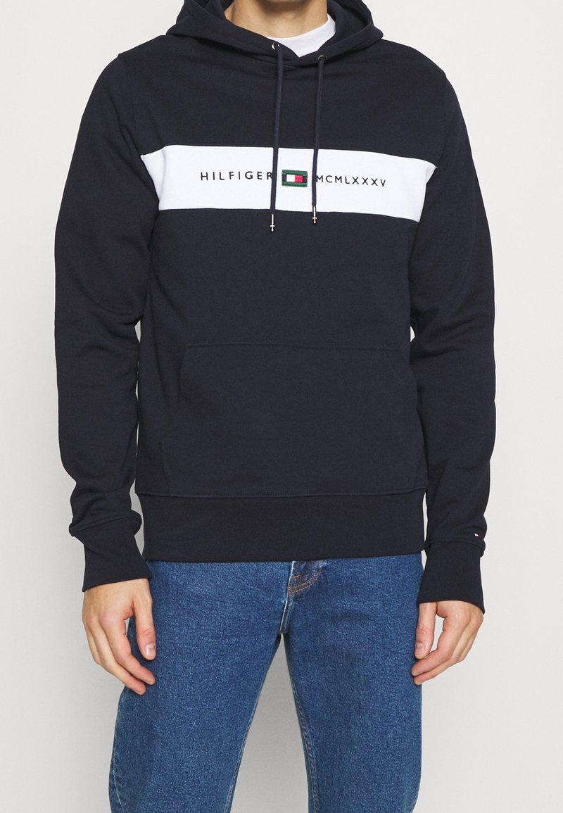 Homme portant un sweat à capuche noir avec une bande blanche sur la poitrine affichant le texte et le logo "Hilfiger MCMLXXXV", associé à un jean bleu.
