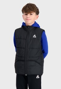 Gilet rembourré noir avec un col haut, fermeture éclair et deux poches avant, porté par-dessus un sweat à capuche bleu, comporte un logo sur la poitrine gauche.