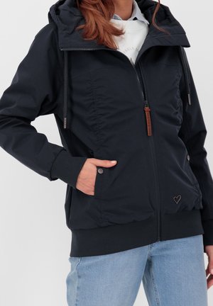 Kobieta ubrana w czarną kurtkę na zamek z kapturem, z prążkowanymi mankietami, z ręką w kieszeni, w połączeniu z jasnoniebieskimi dżinsami i białą koszulką pod spodem.