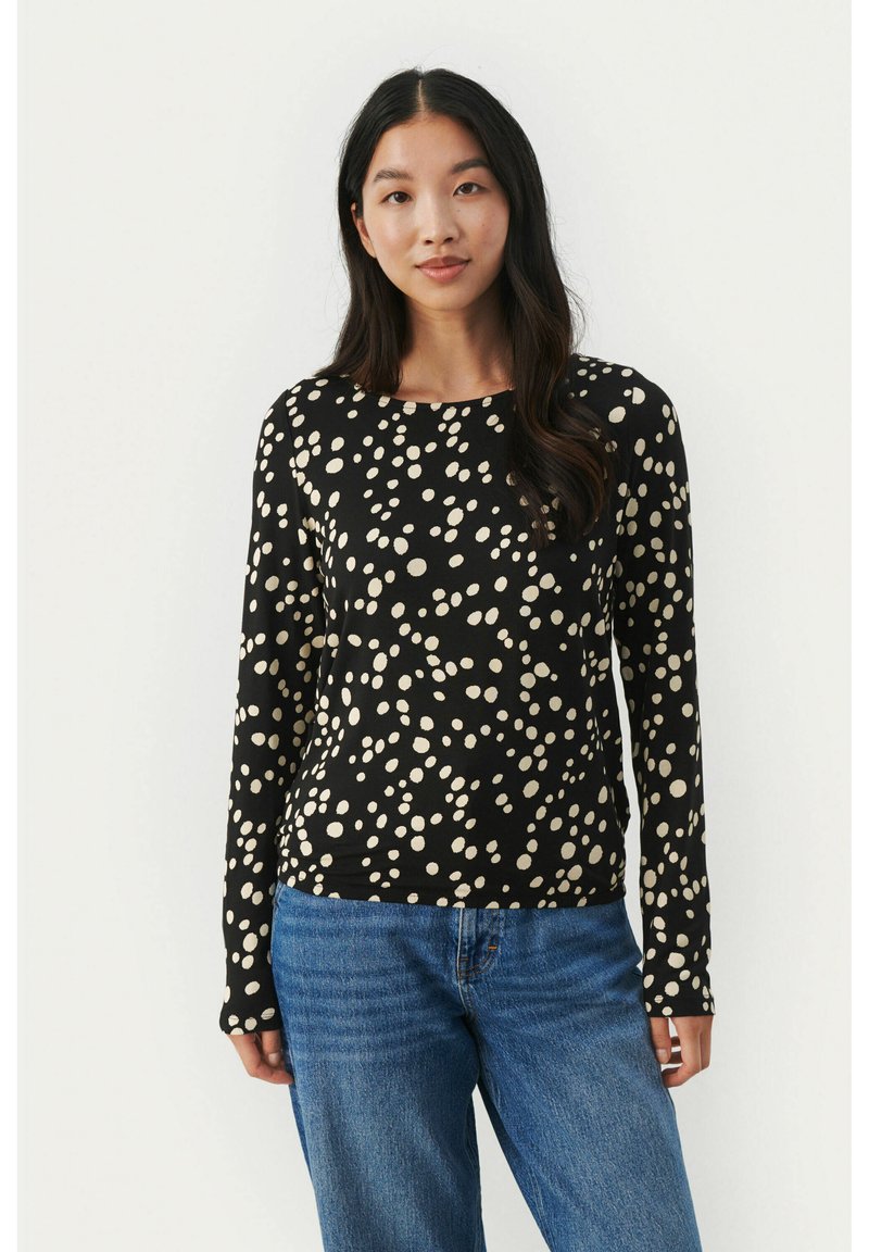 Black long-sleeve top featuring a scattered cream dot pattern, round neckline, and soft, stretchable fabric. Paired with blue denim jeans.