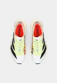 Valkoiset ja mustat urheilukengät kirkkaankeltaisilla yksityiskohdilla, joissa on verkkopäällinen, sävyyn sopivat nauhat sekä sisävuorissa punainen "TERREX"-logo.