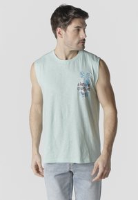 Hombre con camiseta sin mangas azul claro con gráfico de palmera y texto "Vive bajo el sol", combinado con vaqueros de mezclilla claros, mirando hacia un lado.