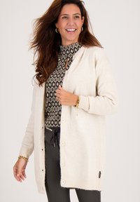 Lichte beige cardigan van zacht materiaal, met een knoopsluiting aan de voorkant, geribde manchetten en een zijlabel. Draagt over een gemusterde top.