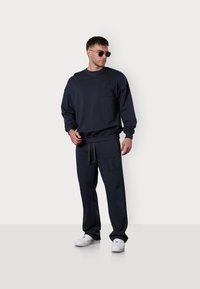 Sweatshirt bleu marine et joggers assortis en tissu doux, avec une poche ornée de logo et une taille à cordon, associés à des baskets blanches.