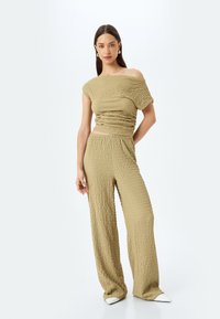 Gebreide, off-shoulder jumpsuit in aardse groene kleur. Textuurstof met een relaxte snit. Wijdlopende pijpen, elastische tailleband, gecombineerd met witte puntige schoenen.