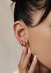 Złote kolczyki w kształcie kółek z polerowanym wykończeniem, noszone na uchu, ukazujące swój okrągły kształt i gładką teksturę. Palce dotykają płatka ucha.