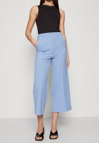 Světle modré široké kalhoty s předními sklady a bočními kapsami, v kombinaci s černým topem bez rukávů a černými sandály na otevřenou špičku.