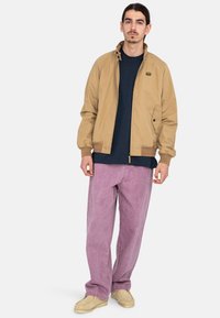 Beige bomber jacket with a high collar, navy t-shirt underneath, and light pink wide-leg pants. Adorned with a small embroidered logo.