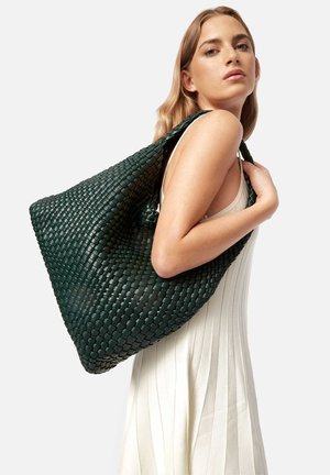 Groene geweven handtas met een textuurafwerking, met een slouchy ontwerp en brede banden, in contrast met een lichtgekleurde jurk.