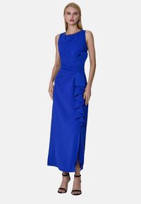 Femme portant une robe maxi sans manches de couleur bleu roi avec des volants asymétriques et des sandales à talons hauts noires, se tenant sur un fond uni.