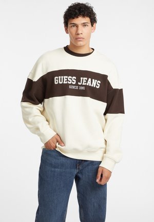 Crewneck sweatshirt i cremefarve og mørkebrun med teksten "GUESS JEANS SINCE 1981". Blødt materiale med ribbede manchetter. Båret med blå denimjeans.