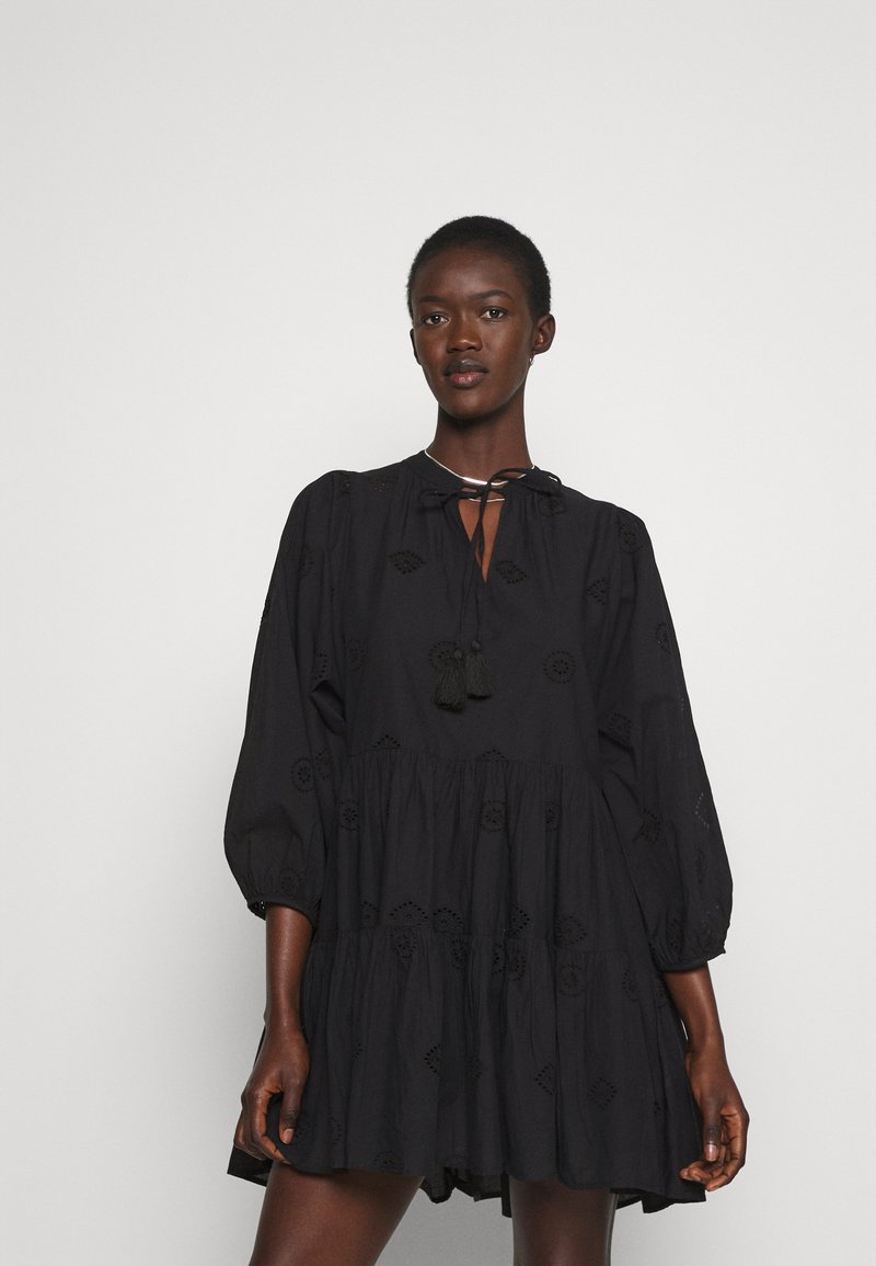 Black dress made of lightweight fabric, featuring a tiered design, balloon sleeves, embroidered details, and a tie neckline with tassels.