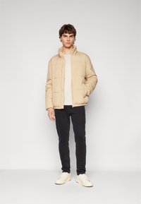 Beige puffer jacket with a high collar, zip closure, and a branded logo on the chest, worn over a light shirt and black pants.