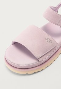 Lys rosa sandal med et mykt suede-overside, bred stropp med UGG-logo og en teksturert gummisåle med kremfarge-akcent.
