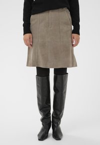 Wildleder-Rock in A-Linie in hellem Taupe, mit vertikalen Nähten und glatter Textur, kombiniert mit kniehohen schwarzen Lederstiefeln.