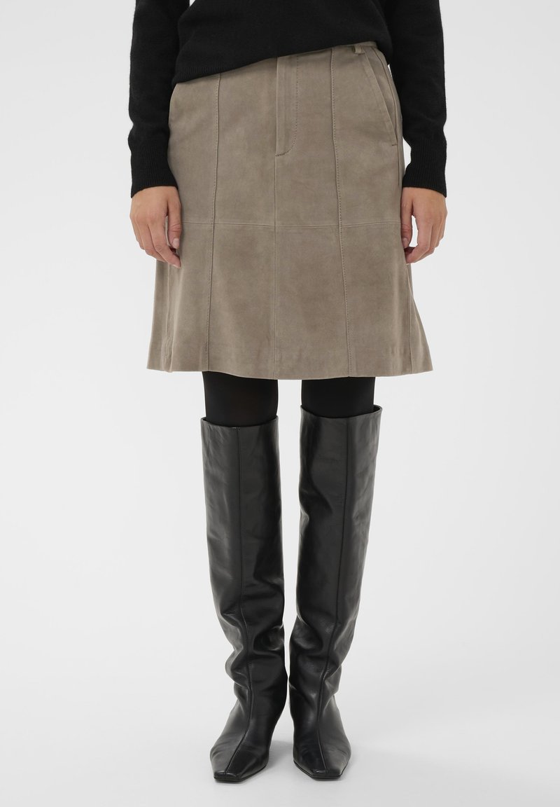 Világos taupe színű velúr A-vonalú szoknya, függőleges varrásokkal és sima textúrával, térdig érő fekete bőrből készült csizmával párosítva.