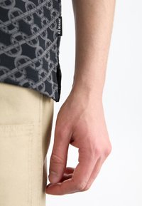 Chemise noire à motifs avec un design en chaîne, fabriquée en tissu avec une étiquette visible sur le côté, associée à un pantalon beige clair.