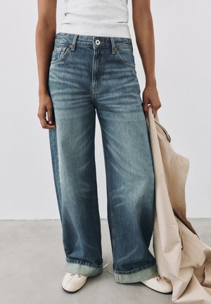 Personne portant un jean bleu à jambes larges et des chaussures blanches, tenant une veste beige dans sa main droite sur un fond uni.
