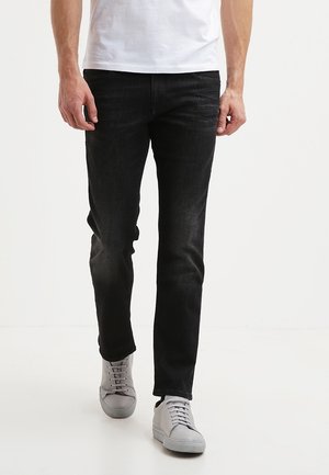 Mand iført slim-fit sorte jeans og lysegrå sneakers, der går fremad mod en ensfarvet hvid baggrund.