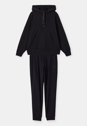 Črna kapuca z sprednjim žepom in vrvico za zategovanje, v kombinaciji z ustreznimi hlačami z elastičnim pasom in stranskimi žepi. Mehka tekstura tkanine.