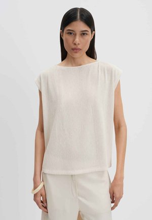 Femme aux cheveux lisses et foncés portant un haut côtelé crème sans manches et un pantalon de couleur claire, debout devant un fond clair uni.