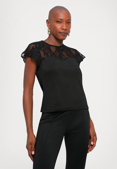 Femme avec la tête rasée portant des boucles d'oreilles en or et un top noir avec des manches en dentelle se tenant devant un fond uni.