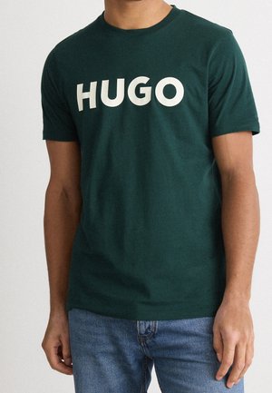 Mies, joka pukeutunut tummanvihreään lyhythihaiseen T-paitaan, jossa on suuri valkoinen "HUGO"-teksti, ja sinisiin farkkuihin, seisoo vaaleaa yksiväristä taustaa vasten.