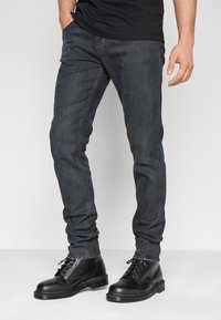 Muž v úzkých tmavě modrých džínách a černých kožených šněrovacích botách stojí před jednoduchým bílým pozadím.