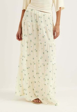 Kobieta w długiej kremowej warstwowej spódnicy z małym niebieskim kwiatowym nadrukiem i beżowych sandałach, stojąca na tle jednolitego jasnego tła.