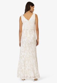 Langes weißes Kleid mit floralem Spitzenoverlay, ärmelloses Design, V-Ausschnitt im Rücken und einem verdeckten Reißverschluss. Sanfte Textur mit kunstvollen Mustern.
