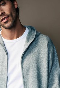 Szara bluza z kapturem zapinana na zamek, wykonana z miękkiego materiału, noszona na białej koszulce z okrągłym dekoltem, charakteryzująca się luźnym krojem i kapturem.